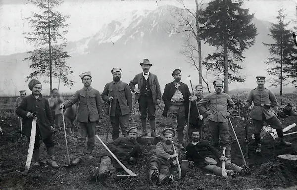 Eine Gruppe russischer Kriegsgefangener mit Holzfällerwerkzeug und ein Wachsoldat vor einer Bergkulisse.