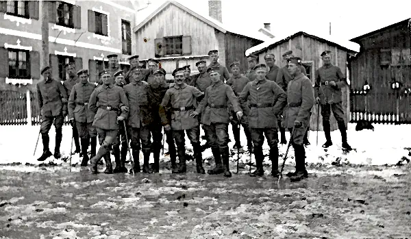 Eine Gruppe deutscher Soldaten in feldgrauen Uniformen und Wickelgamaschen vor winterlichen Holzhütten.